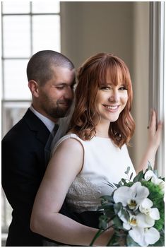 a bride and groom standing next to each other in front of a window with white flowers