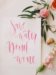 pink watercolor lettering on white paper next to flowers and wine glasses with corks