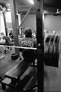 a man squats on a bench in a gym with other people working out behind him