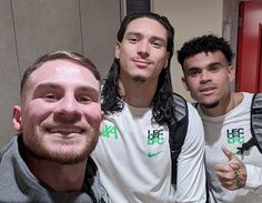 three young men posing for a photo in front of a locker room door, one with long hair and the other with beards