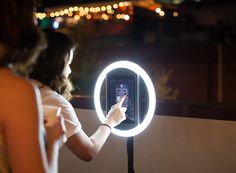 a woman taking a photo with her cell phone in front of a circular light up mirror