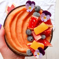 a bowl filled with fruit and flowers on top of a white table next to a person's hand