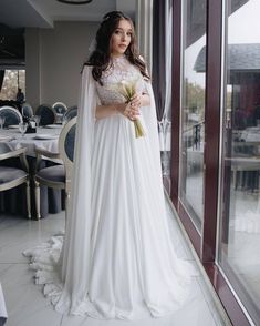 a woman in a white wedding dress standing next to a window holding a flower bouquet