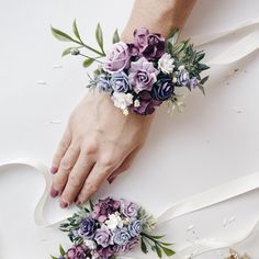 two brides hands with purple and white flowers on their wristbands, one holding the other