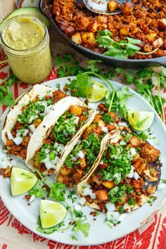 three tacos on a white plate with limes and cilantro next to it