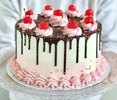 a person holding a cake with chocolate and pink icing on it, topped with cherries