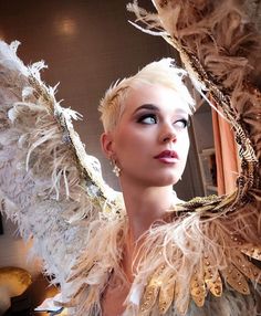 a woman with white hair and feathers on her head is looking into the distance while standing in front of a mirror