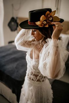 a woman in a white dress and black hat with flowers on her head is standing next to a bed