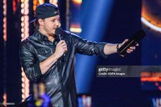 a man in black leather jacket and cap holding a microphone on stage with lights behind him