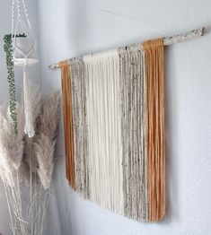 a wall hanging made out of yarn and wood sticks with some plants in the corner