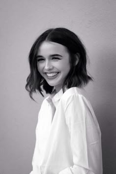 a young woman smiling and wearing a white shirt with her hands in her pockets, standing against a gray wall