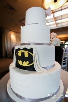 a three tiered white cake with a batman symbol on the top and silver trim