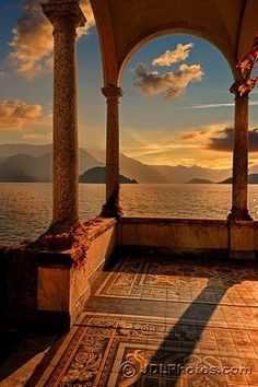 the sun is setting over an ocean with pillars and arches on it, as seen from across the water