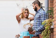 a man and woman standing next to each other by a brick wall with plants on it