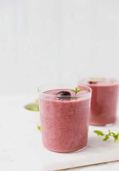 two glasses filled with smoothie sitting on top of a white counter next to green leaves