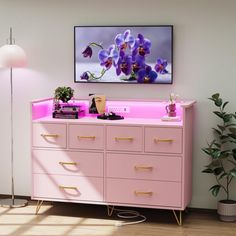 a pink dresser with purple flowers on it in front of a large framed painting and potted plant