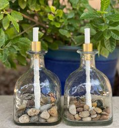 two glass bottles with candles inside sitting next to each other on a table near a potted plant