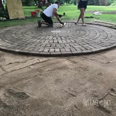 two people working on a brick patio in the middle of a yard with grass and trees