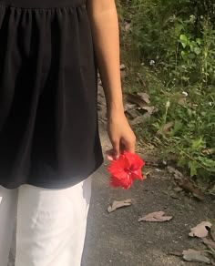 a woman holding a red flower in her hand while walking down a path next to some bushes