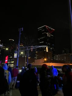 a group of people standing on the side of a road at night with tall buildings in the background