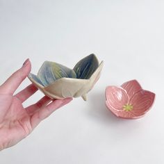 a hand holding a small ceramic bowl next to a tiny pink flower shaped object on a white surface