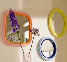 a vase filled with purple flowers sitting next to two colorful mirrors on the side of a wall