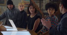 a group of people sitting around a wooden table with papers on it and one person standing in front of them