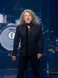 a man with long hair standing on stage in front of a microphone and drum set