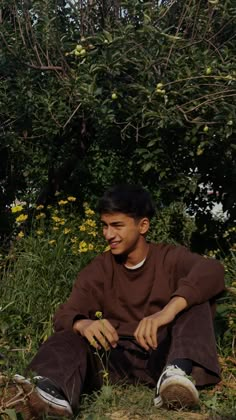 a man sitting on the ground in front of some bushes and flowers with his hands together