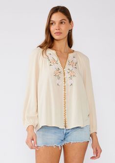 [Color: Natural/Pink] A front facing image of a brunette model wearing a cream colored bohemian blouse with floral embroidered details. With long raglan sleeves Casual V-neck Blouse With Embroidered Neckline, Bohemian V-neck Blouse With Embroidered Hem, Casual V-neck Blouse With Embroidered Sleeves, Spring Floral Embroidered V-neck Top, Fall V-neck Blouse With Embroidered Sleeves, Embroidered V-neck Blouse For Fall, Feminine Spring Tops With Split Neck, Casual V-neck Blouse With Embroidered Hem, Folk Style V-neck Blouse With Embroidered Neckline