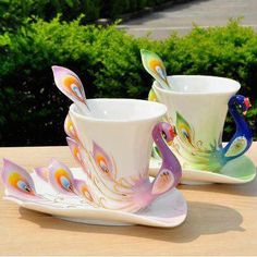 two coffee cups and saucers decorated with peacocks on them sitting on a table