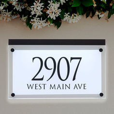 a white and black sign hanging from the side of a building with flowers in the background