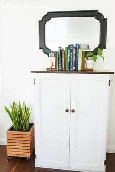 a white cabinet with books on top and a mirror above it next to a potted plant