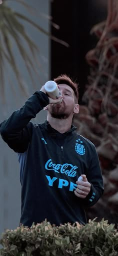 a man drinking from a bottle while standing in front of a plant and looking up at the sky