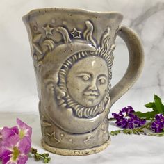 a ceramic mug with a sun face on it next to some purple flowers and leaves