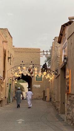 two men walking down an alley way with lights hanging from the ceiling and decorations above them