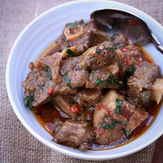 a white bowl filled with meat and vegetables on top of a wooden table next to a spoon