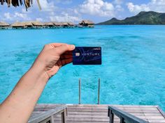 a hand holding up a credit card in front of the ocean and over water huts