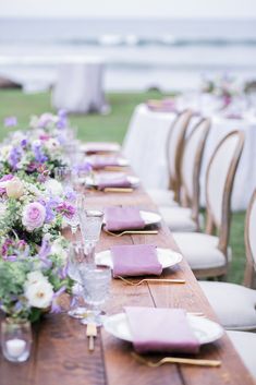 a long table with purple napkins and flowers on it is set for an outdoor dinner