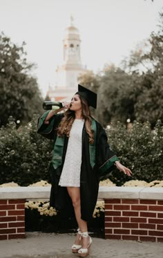 a woman in a graduation gown drinking from a bottle while standing on a brick wall