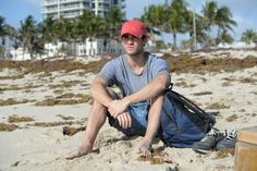 a man sitting on the beach with his surfboard in front of him and palm trees behind him