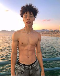 a shirtless young man standing in front of the ocean at sunset with his hands on his hips