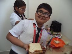 a boy and girl standing in front of a table with food on it, one holding a sandwich