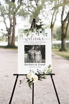 a welcome sign with flowers on it in the middle of a park area for an outdoor wedding