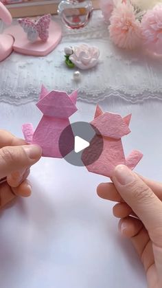 two hands holding origami pigs in front of a table with pink flowers on it