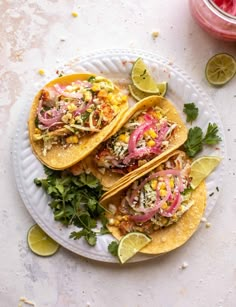 three tacos on a plate with limes and cilantro next to it