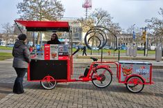 a woman standing next to a red bike with a cart on the back that is attached to it