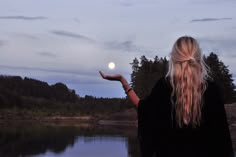 a woman with long blonde hair is holding out her hand in front of the water