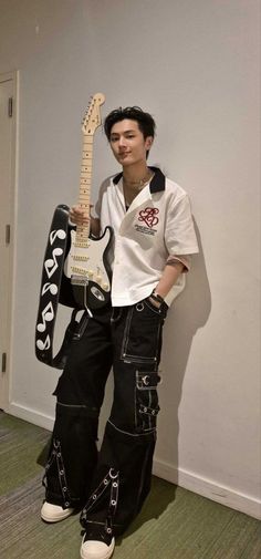 a man in black pants and white shirt holding an electric guitar while standing next to a wall