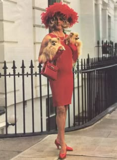 a woman in a red dress and matching hat holding two small dogs while walking down the street